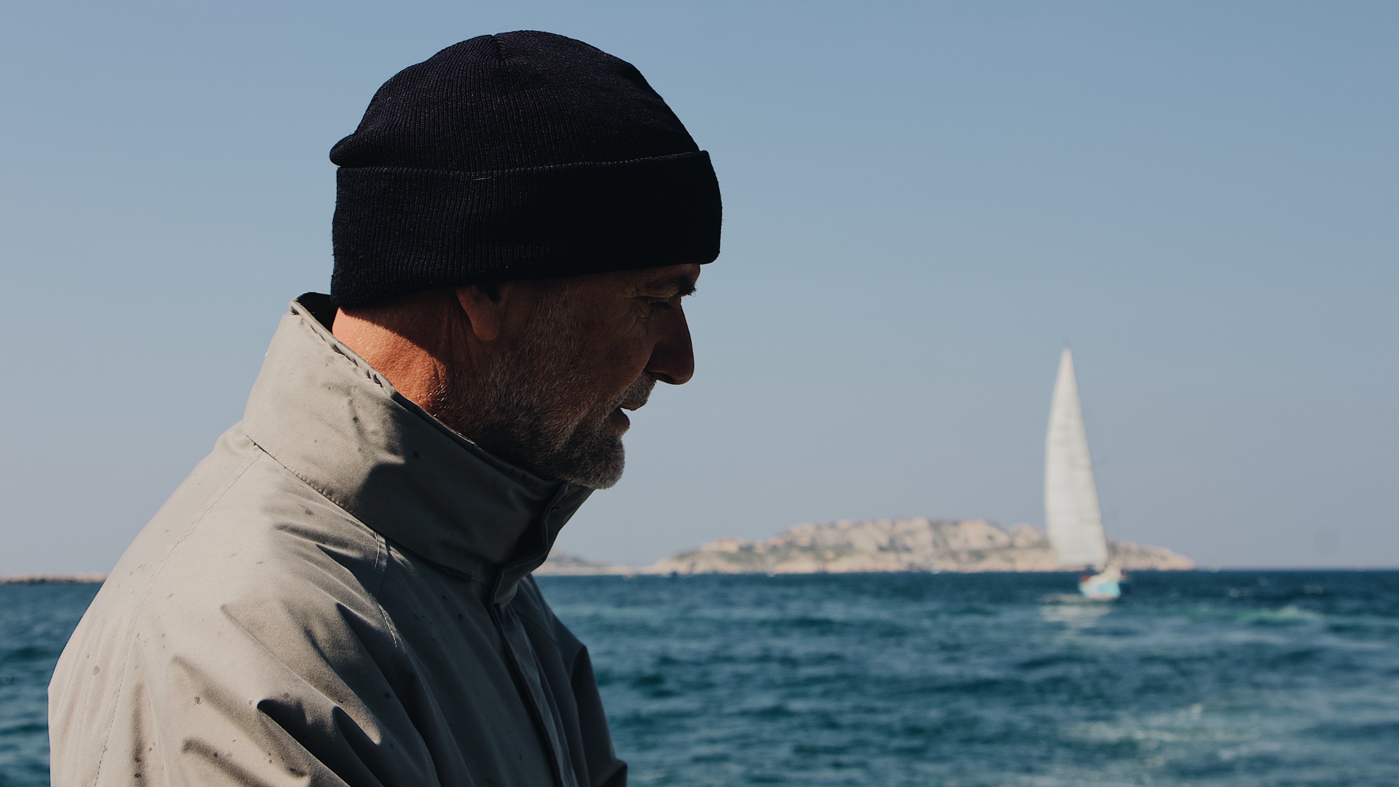 Mann med lue med seilbåt, sjø og en øy i bakhgrunnen. Foto av Mael BALLAND på unsplash.com/@mael_bld